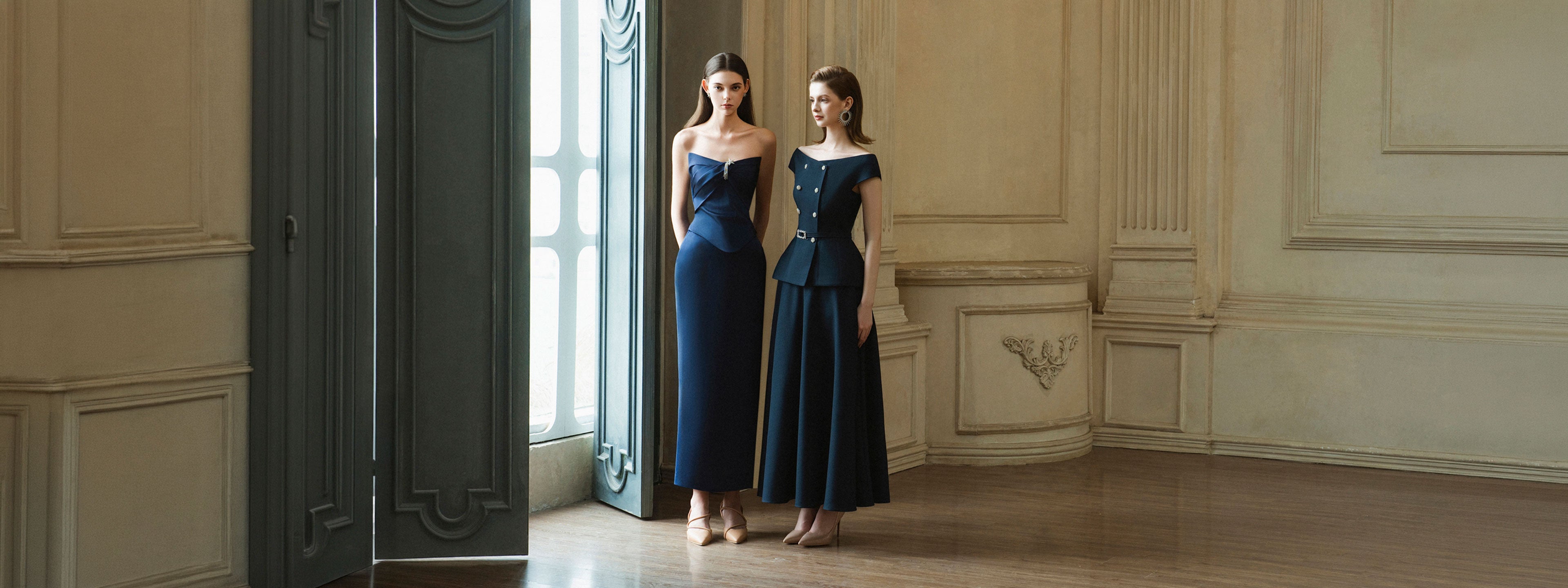Two models in navy off-the-shoulder maxi dresses from DeTHEIA's De Nona Cometa collection, in an elegant classical interior